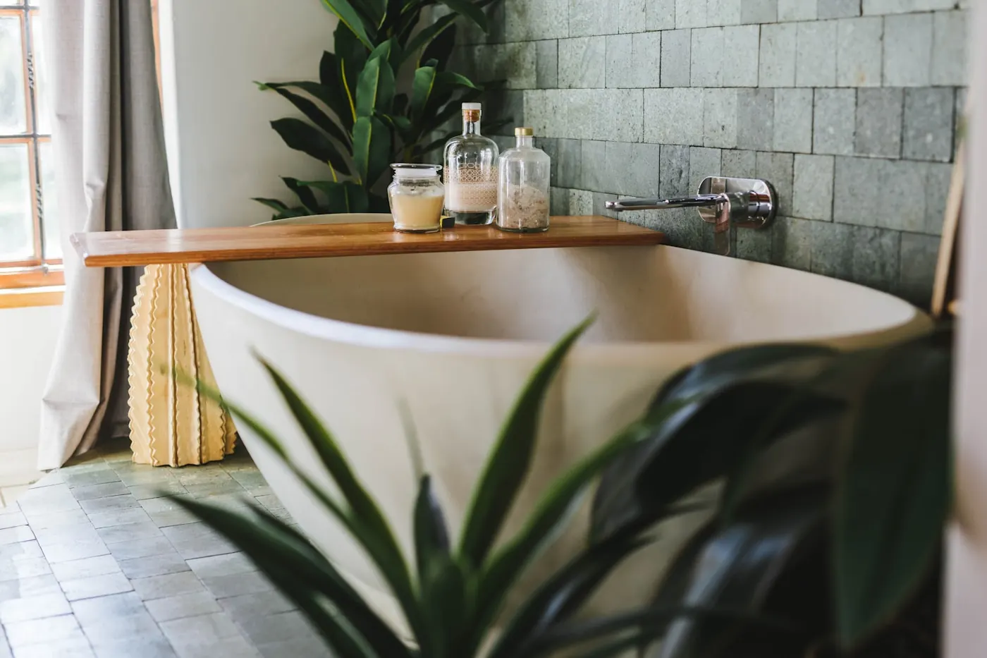 Freestanding oval bathtub with a wooden bath tray against a stone-tiled wall in a naturally lit bathroom